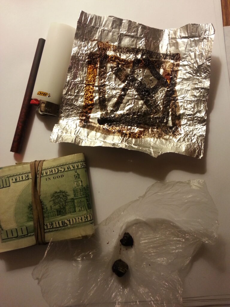 Image of Heroin Paraphernalia. I white cigarette lighter, a cut black straw (tooter) small square of aluminum foil with black and amber burn marks on it, a small baggie showing a dark black substance, and a wad of money with a rubber band around it.