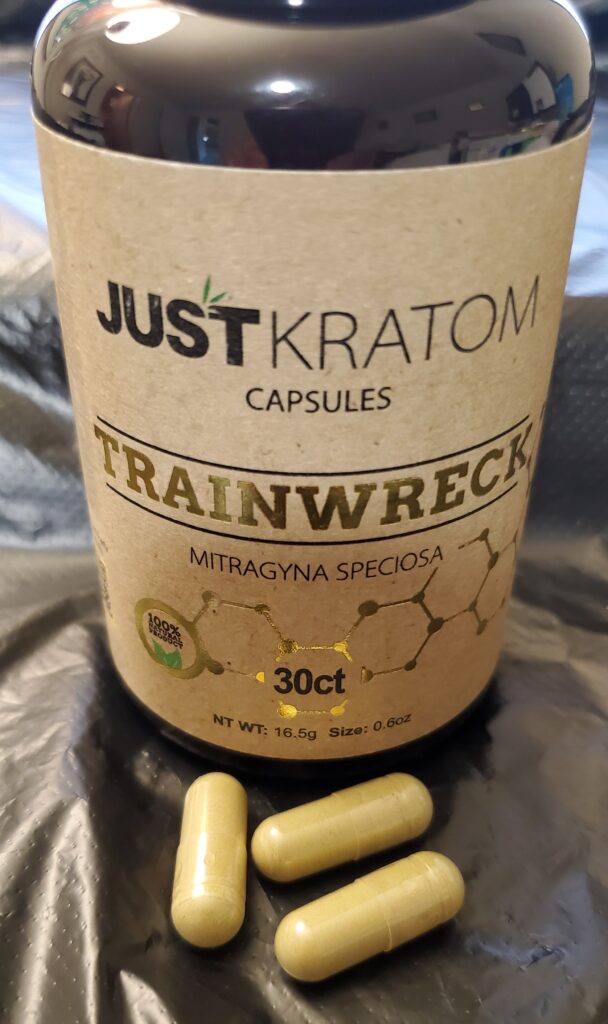 Image is a brown bottle looking container with a tan label wrapped around it. In from of the bottle are 3 yellow looking capsules. The writing on the label says JUST KRATOM on top with CAPSULES below it. Under that is the word TRAINWRECK with a black line above and below the word. Below that is Mitragyna Speciosa with a chemical structure under that and 30 ct in the middle.