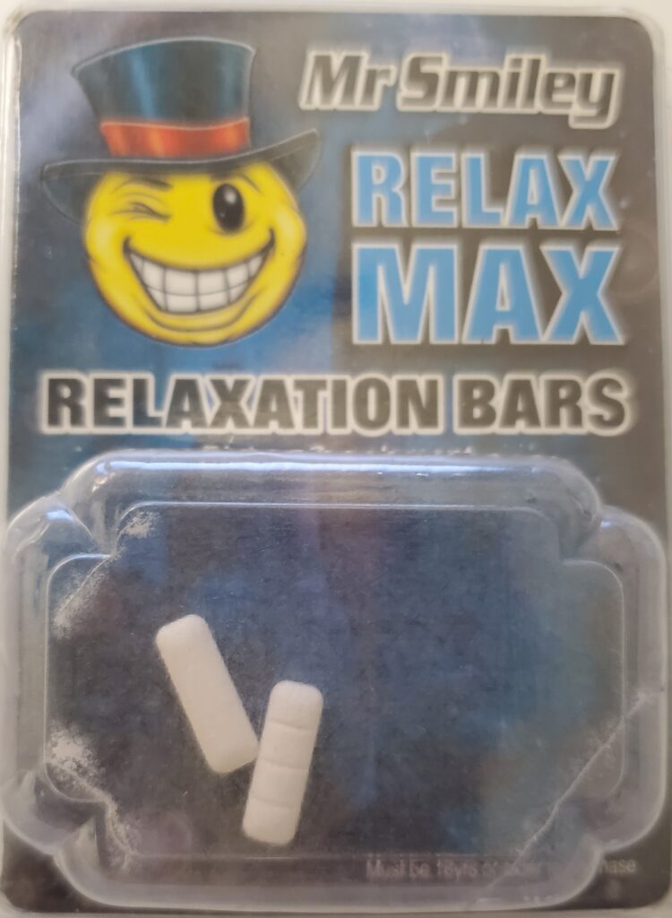 Image is of a rectangle packet with an image of a smilely face winking and wearing a black top hat with a red band around the crown above the brim. Verbiage on top right says, "Mr. Smiley Relax Max relaxation bars." The bottom has a rectangular puffed out plastic area that contains 2 white, long, thin rectangles with 3 lines across them. The look like 4 small squares stacked on top of each other. They resemble prescription Xanax pills but contain Phenazepam, not Alprazolam.