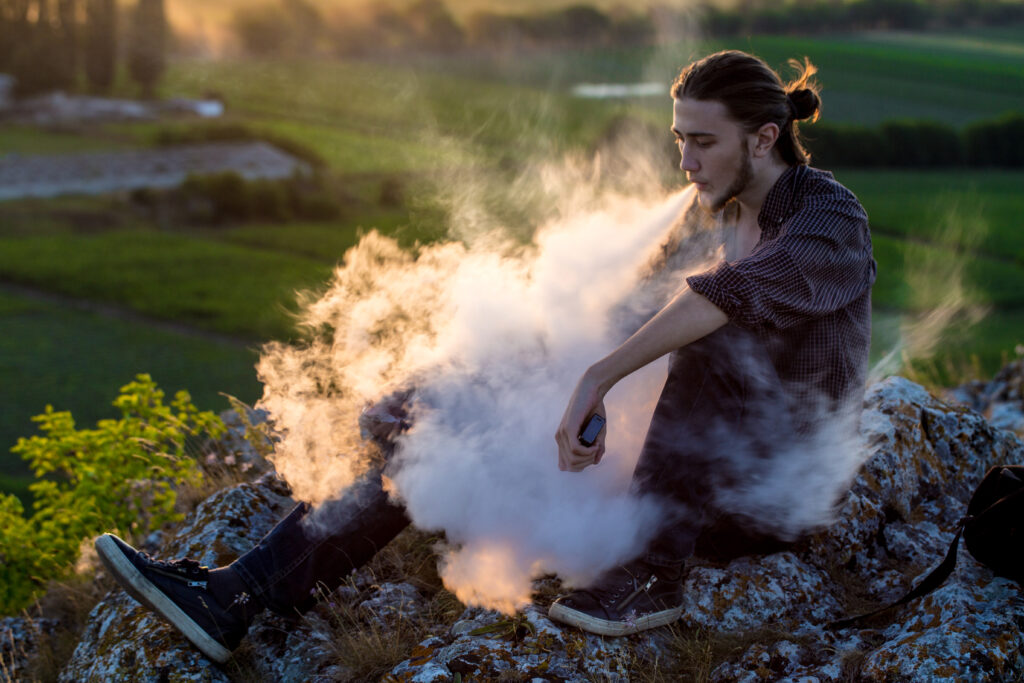 Handsome young man sitting on a rock and smoking electronic cigarette outdoors.