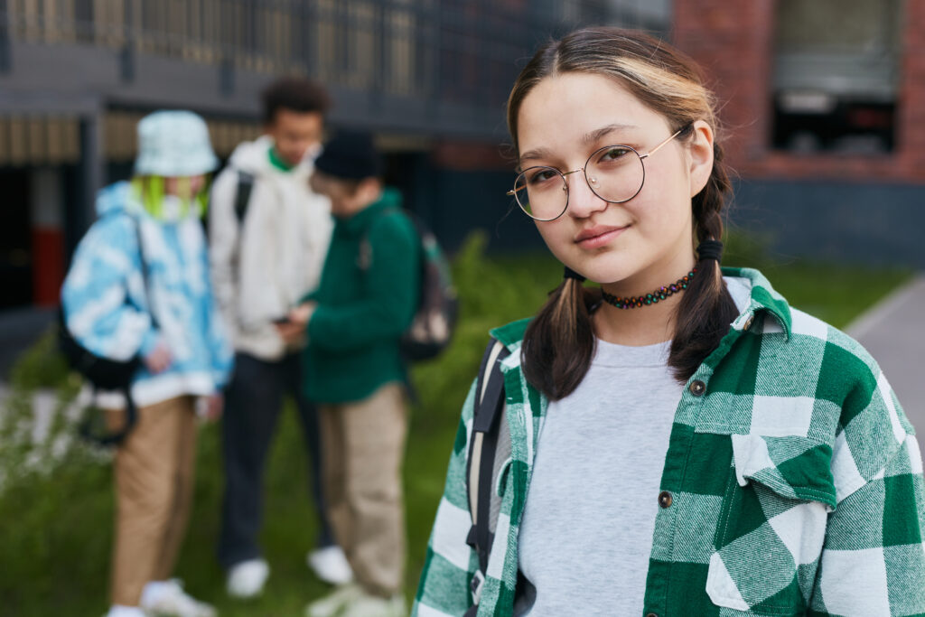 Young schoolgirl in eyeglasses and checked shirt looking at the camera while standing outside with her classmates in background.